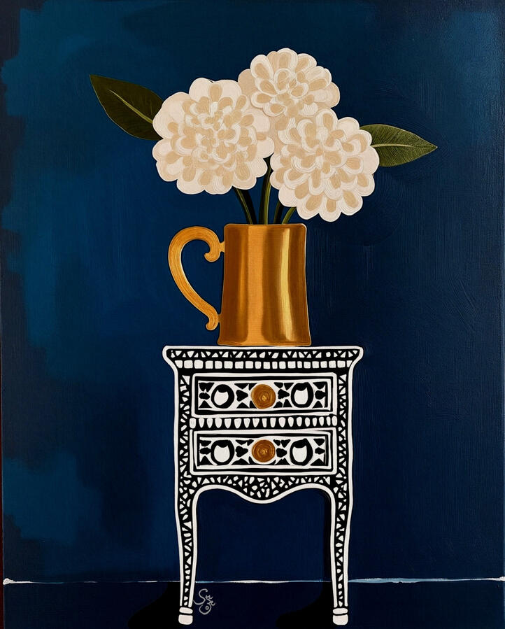Cream hydrangea flowers in a matter of gold jar sitting atop a white patterned black vintage inlaid cabinet. Deep blue background.