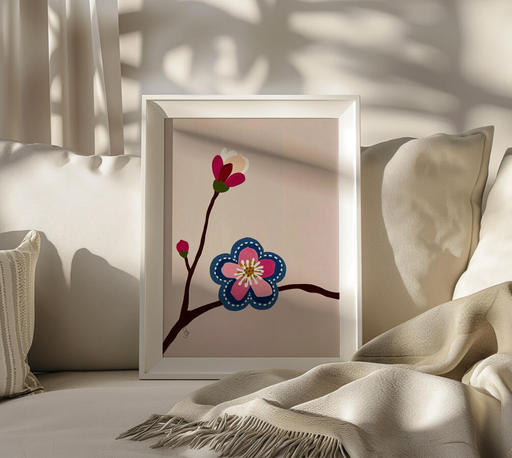 A white sleek picture frame resting on cosy couch, containing an illustration of a pink blossom with stitched denim edges. Hover Text Reads. “Visit Goodmoodprints to take it home"