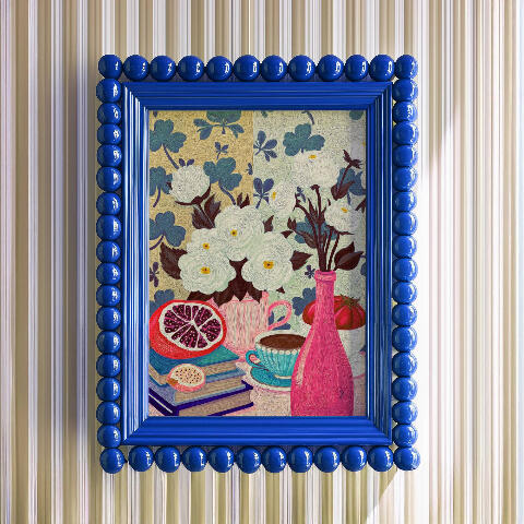 Framed mockup of a colorful still life painting with white flowers, clover leaves, books, fruit, and a pink bottle displayed in a bright blue decorative frame.